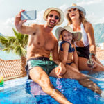 Father, wife and daughts in pool on vacation taking selfie in front of mounatains and the sea.