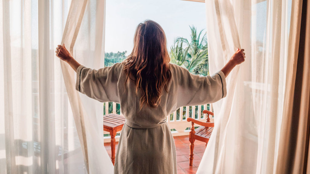 woman opening the curtains to a beautiful tropical view from her hotel room, which she saved on booking through hotels.com black friday