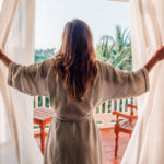 woman opening the curtains to a beautiful tropical view from her hotel room, which she saved on booking through hotels.com black friday